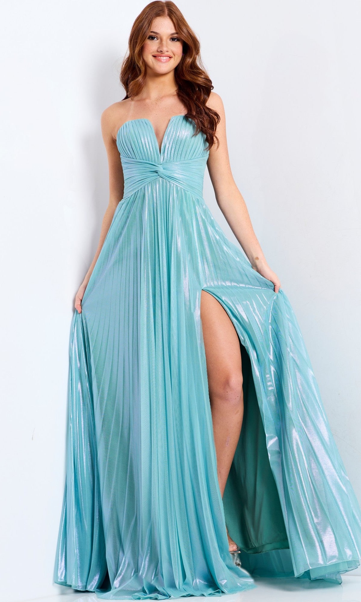 A woman wears the Jovani 39023 long prom dress in mint, featuring a strapless shimmery pleated bodice, high slit, and flowing fabric, standing against a plain light background.
